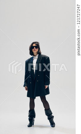 Full-length portrait of stylish young woman wearing black oversized jacket, sunglasses and heeled boots, standing in a confident pose against studio background Full-length portrait of stylish young woman wearing black oversized jacket, sunglasses and heeled boots, standing in a confident pose against studio background 121727247