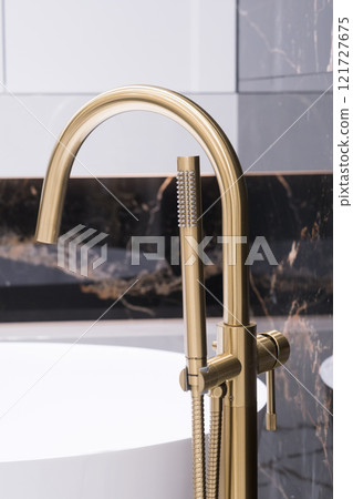 Long stylish Gold faucet on the floor near white bathtub on granite wall background. Modern bathroom interior 121727675