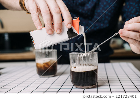 Pouring milk into iced coffee in a glass. Home barista concept, minimal tabletop aesthetic, beverage preparation. 121728401