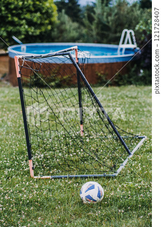 A backyard scene featuring a small soccer goal with a ball on a green lawn, set beside an above-ground swimming pool surrounded by trees. A backyard scene featuring a small soccer goal with a ball on a green lawn, set beside an above-ground swimming pool surrounded by trees. 121728407