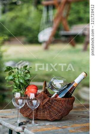 Rustic wicker basket with wine, glasses, basil, and tomatoes on a wooden table in a serene outdoor setting. Farm-to-Table Living concept. Rustic wicker basket with wine, glasses, basil, and tomatoes on a wooden table in a serene outdoor setting. Farm-to-Table Living concept. 121728412