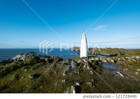 Irish landmark. A white navigational marker is visible against a backdrop of a rocky Irish coastline and expansive blue sea. The sky is cloudless. Irish landmark. A white navigational marker is visible against a backdrop of a rocky Irish coastline and expansive blue sea. The sky is cloudless. 121728490