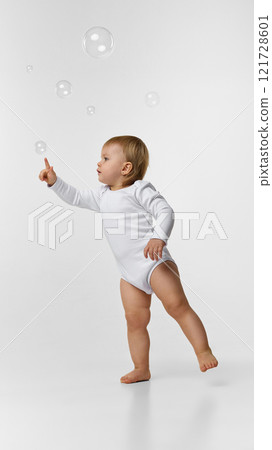 Baby girl, toddler in white bodysuit reaching for floating bubbles with interest and curiosity against white studio background 121728601