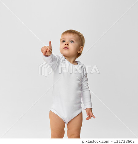 Baby girl, toddler in white bodysuit looking and pointing upwards with interest and curiosity against white studio background 121728602