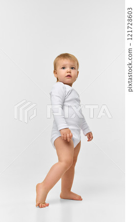 Baby girl, toddler in white bodysuit standing with cute expression, looking with attention against white studio background 121728603
