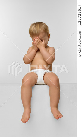 A baby girl, toddler in a diaper sitting on a white cube, and playfully covering face with hands against studio background 121728617