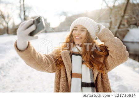 A young woman in a snowy park with a phone in her hands communicates via video and takes a selfie. A young woman in a snowy park with a phone in her hands communicates via video and takes a selfie. 121730614