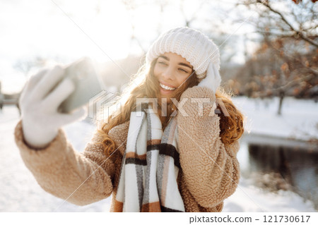 A young woman in a snowy park with a phone in her hands communicates via video and takes a selfie. A young woman in a snowy park with a phone in her hands communicates via video and takes a selfie. 121730617