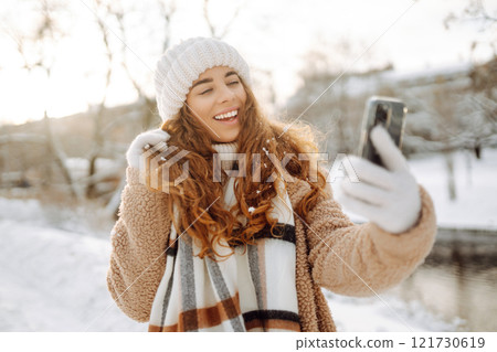 A young woman in a snowy park with a phone in her hands communicates via video and takes a selfie. 121730619