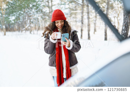 A young traveler in a red hat sits in the trunk of a car and drinks a hot drink from a thermos. 121730885