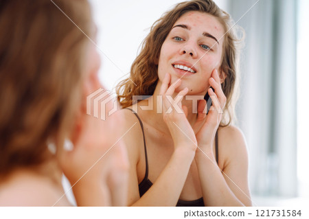 Beautiful woman touches her inflammation on her face while looking in the mirror in a bright room. Beautiful woman touches her inflammation on her face while looking in the mirror in a bright room. 121731584
