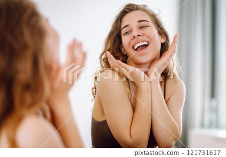 Beautiful woman touches her inflammation on her face while looking in the mirror in a bright room. Beautiful woman touches her inflammation on her face while looking in the mirror in a bright room. 121731617