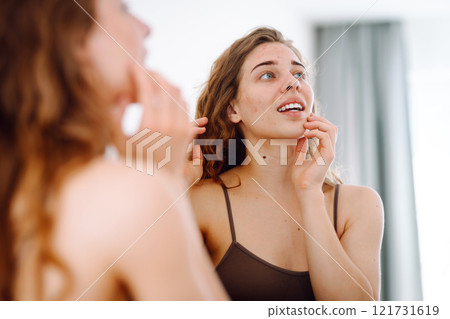Beautiful woman touches her inflammation on her face while looking in the mirror in a bright room. Beautiful woman touches her inflammation on her face while looking in the mirror in a bright room. 121731619
