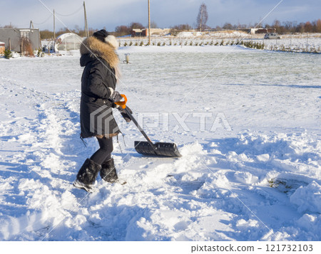 Woman wearing heavy winter gear shoveling snow in bright sunlight, clearing path through snowy landscape with determined movement 121732103