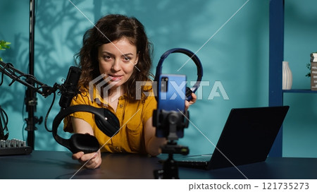 Audio engineer recording video for internet channel in dimly lit home studio, presenting pairs of gaming headphones to audience. Woman showing viewers music listening device, camera A 121735273