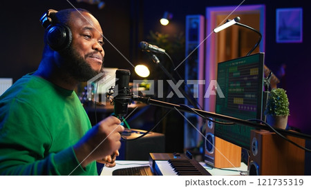 Confident artist singer performing a song on microphone in home studio, recording his audio files and manipulating raw sound on professional editing software. Music production. Camera B. 121735319