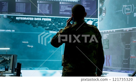Military man answers phone call in a mission control center, working with advanced technology and satellite imagery to track potential threats and real time data. Intelligence activities. Camera A. 121735320