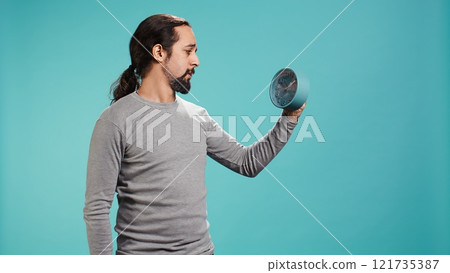 Portrait of worried man looking at alarm clock, stressed after not hearing ring and waking up late in the morning. Anxious person running out of time, isolated over studio background, camera B Portrait of worried man looking at alarm clock, stressed after not hearing ring and waking up late in the morning. Anxious person running out of time, isolated over studio background, camera B 121735387