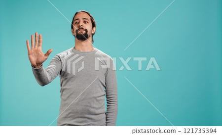 Assertive man doing stop hand gesture sign, studio backdrop. Annoyed person doing firm halt sign gesturing, wishing to end concept, isolated over blue studio background, camera B 121735394