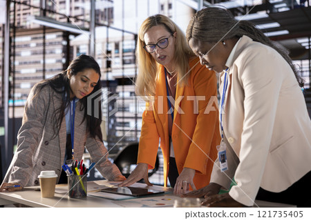 Diverse team of empowered women executives discussing corporate strategy and business growth. Examining analytics to obtain funds for projects, showcasing leadership skills and gender equality. 121735405