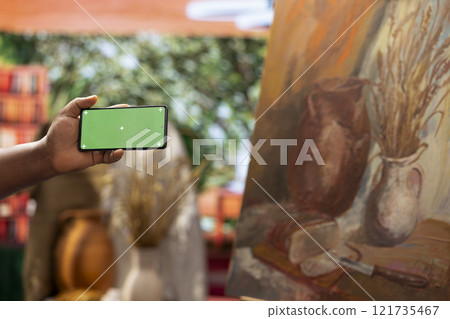 Young artist looking at isolated chroma key display in his painting atelier, using device next to canvas with a colorful masterpiece. Painter creating a rustic artwork for the gallery exhibition. 121735467