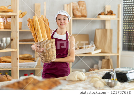 Young girl baker are standing near product range in bakery kitchen with basket loaf of baguettes Young girl baker are standing near product range in bakery kitchen with basket loaf of baguettes 121737688