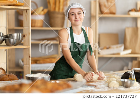 Teen girl works in bakery as baker, kneads dough, works with flour. 121737727