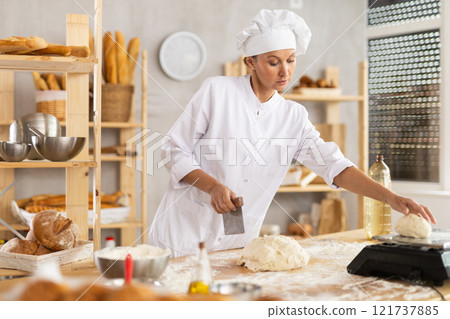 Baker woman cuts small pieces of dough from large lump and puts them on scales. 121737885