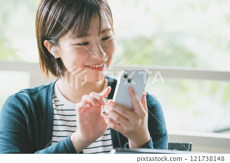 一位年輕女子戴著無線耳機，在鬱鬱蔥蔥的陽台上操作智慧型手機 121738148