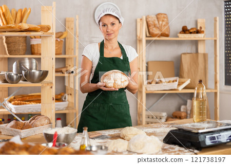 Young woman professional baker in apron standing at work table, posing with baked bread in bakery kitchen 121738197
