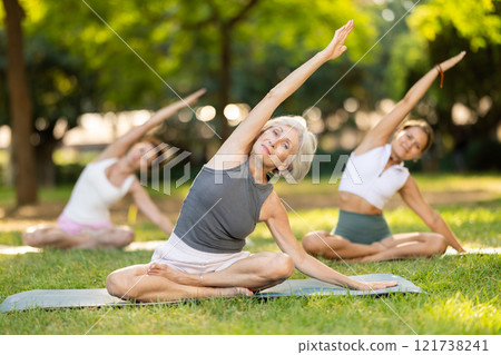 Sporty old lady sitting on sports mat and making yoga meditation in lotus pose and hands up with group of women together in green park at daytime 121738241