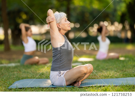 Sporty old lady sitting on sports mat and making yoga meditation in lotus pose and hands up with group of women together in green park at daytime 121738253