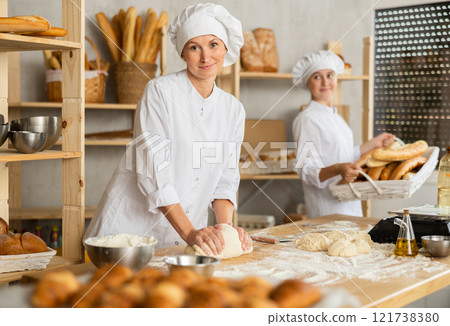 Two professional female baker in uniform working in bakehouse. Adult woman kneading dough and young girl arranging freshly baked bakery products Two professional female baker in uniform working in bakehouse. Adult woman kneading dough and young girl arranging freshly baked bakery products 121738380