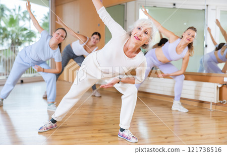 Mature female coach dancing and practicing new movements with group of young women in gym. Dance and wellbeing concept Mature female coach dancing and practicing new movements with group of young women in gym. Dance and wellbeing concept 121738516