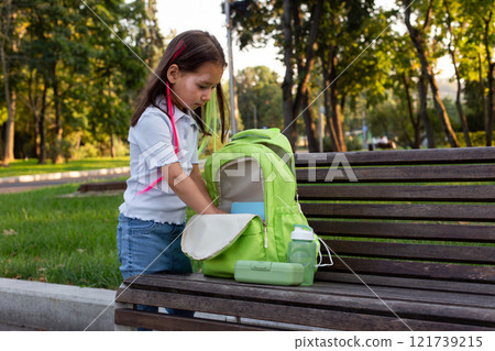 Young girl organizing backpack on park bench in bright, sunny morning 121739215