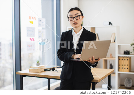 Portrait of young Asian businesswoman standing with laptop in office environment. She wears black suit and glasses, conveying professionalism and confidence. Bright modern workspace with large windows 121742266