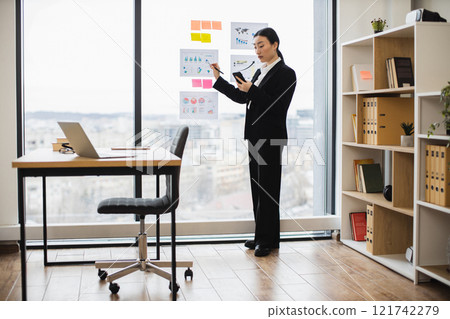 Asian businesswoman in formal attire stands by window analyzing charts on glass wall in contemporary office. Professional environment and thoughtful expression capture focus and dedication. Asian businesswoman in formal attire stands by window analyzing charts on glass wall in contemporary office. Professional environment and thoughtful expression capture focus and dedication. 121742279