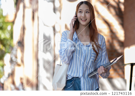 Young woman smiling while talking on the phone, holding a tablet and tote bag outdoors on a sunny day. Young woman smiling while talking on the phone, holding a tablet and tote bag outdoors on a sunny day. 121743116