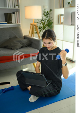Young asian woman lifting a light dumbbell and using digital tablet on yoga mat Young asian woman lifting a light dumbbell and using digital tablet on yoga mat 121744832