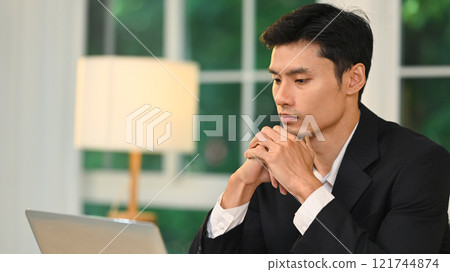 Thoughtful millennial entrepreneur with his hands clasped under his chin working on laptop 121744874