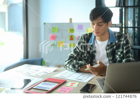 Smiling male UX designer working on app design ideas with detailed sketches spread across the desk 121744876