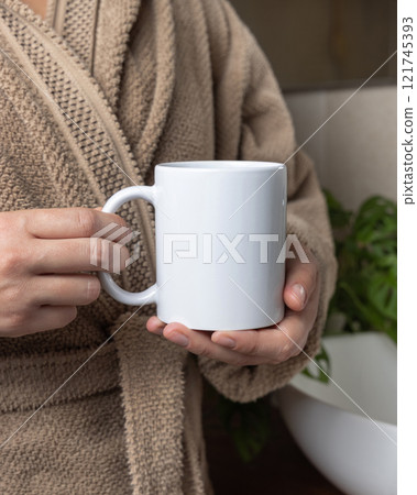 Woman in brown bathrobe holding white blank coffee mug in bathroom closeup,  mockup 121745393