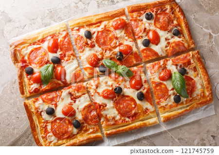 Savory open pie made of puff pastry with pepperoni sausage, olives, mozzarella and tomato sauce close-up on parchment. horizontal top view Savory open pie made of puff pastry with pepperoni sausage, olives, mozzarella and tomato sauce close-up on parchment. horizontal top view 121745759