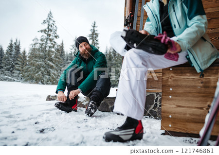 Couple preparing for cross country skiing in winter nature, putting boots on. Sitting in front cabin. Couple preparing for cross country skiing in winter nature, putting boots on. Sitting in front cabin. 121746861