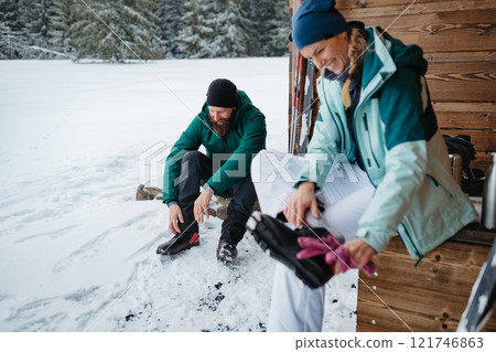 Couple preparing for cross country skiing in winter nature, putting boots on. Sitting in front cabin. Couple preparing for cross country skiing in winter nature, putting boots on. Sitting in front cabin. 121746863