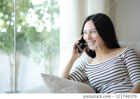 Happy asian woman talking on phone looking through a window 121748379