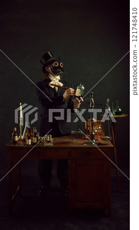 Plague doctor working under candlelight in vintage laboratory with antique medical tolls against dark mystery background. 121748410