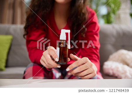 Cropped photo of woman holds brown glass spray bottle with white nozzle while sitting on sofa in living room. 121748439