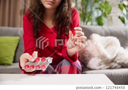 Woman in red pajamas sitting on sofa, holding lozenge and blister pack of red tablets in her hands. 121748450
