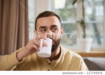Healthy young man drinking hot mineralizing, vitaminizing drink sitting on sofa in cozy living room at home. 121748476
